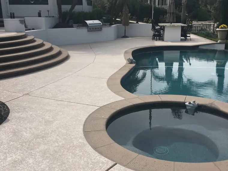Backyard pool and round spa with a gray textured deck and outdoor kitchen behind