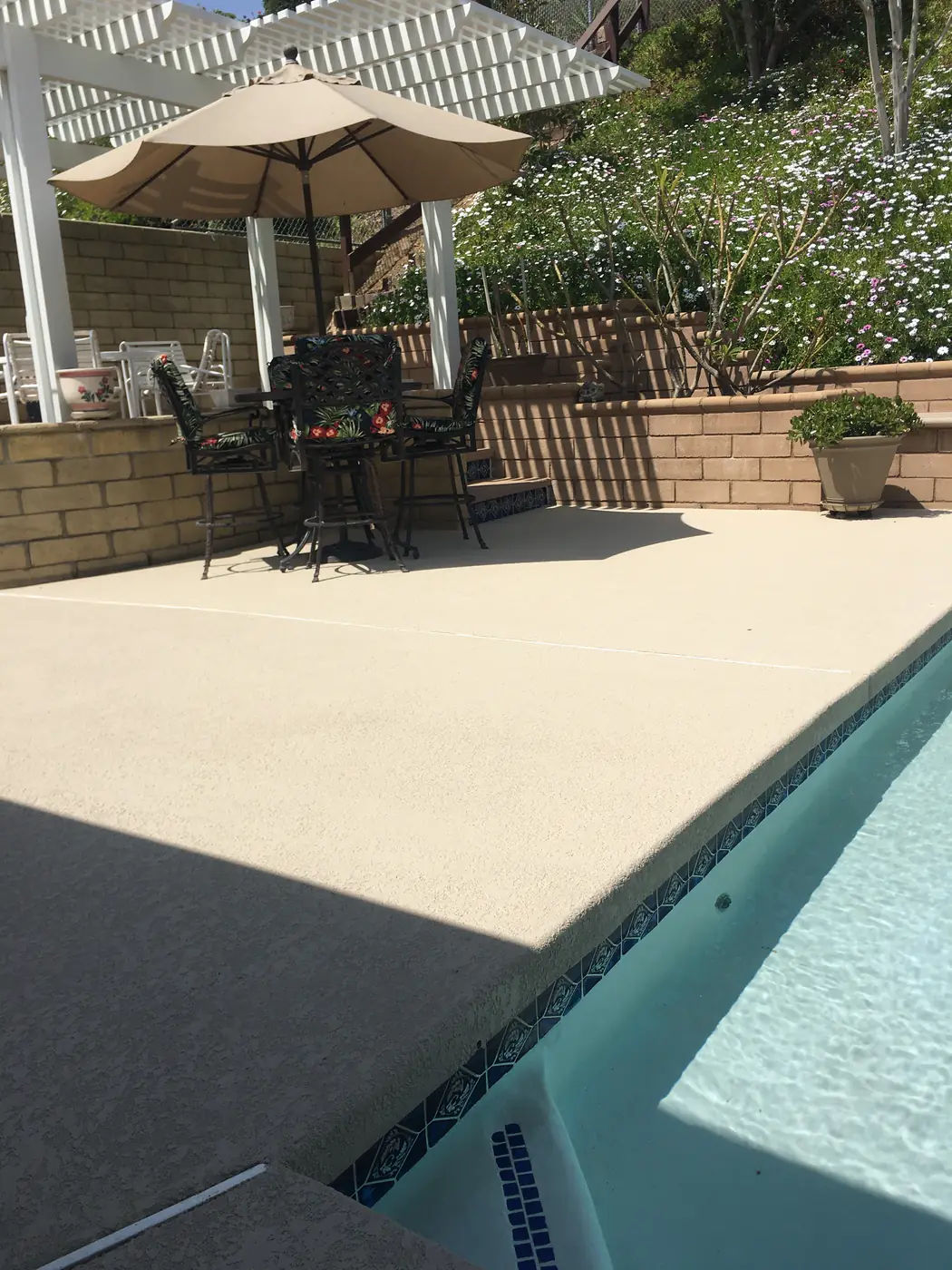 Patio furniture and umbrellas on a cream pool deck beside a backyard pool