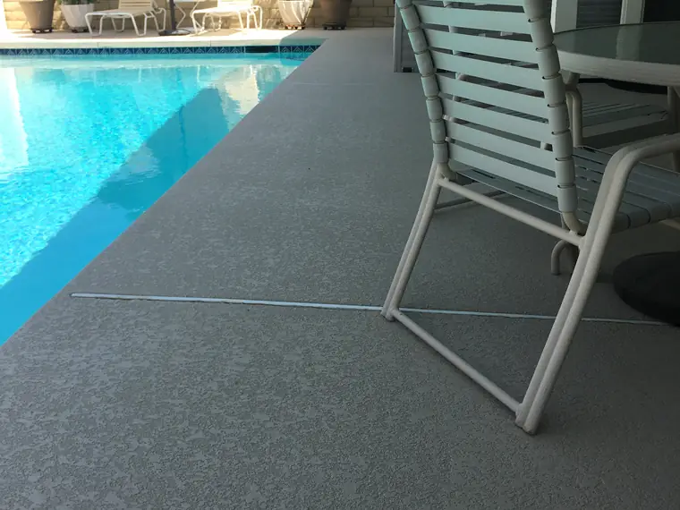 Poolside chairs on a gray textured deck beside clear blue water
