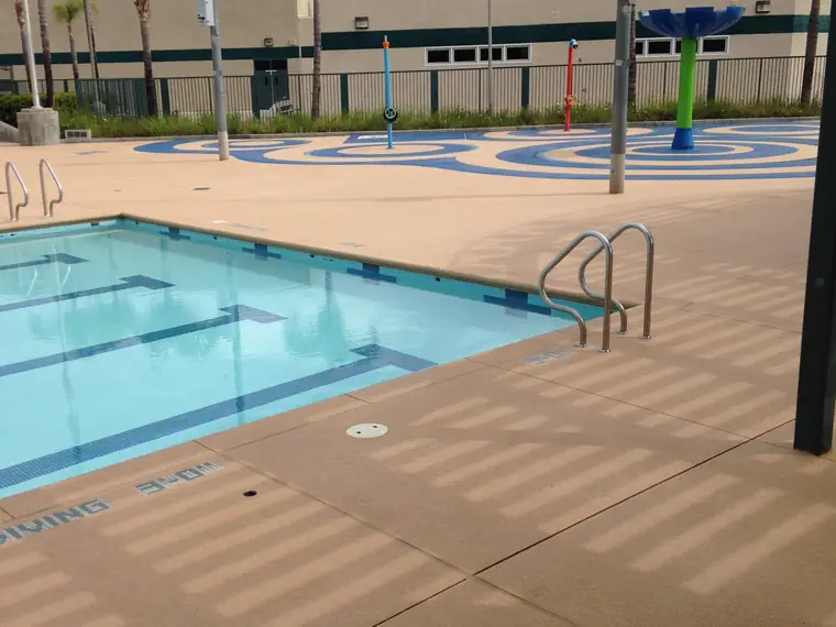 Community pool and splash pad with a tan textured deck and palm trees